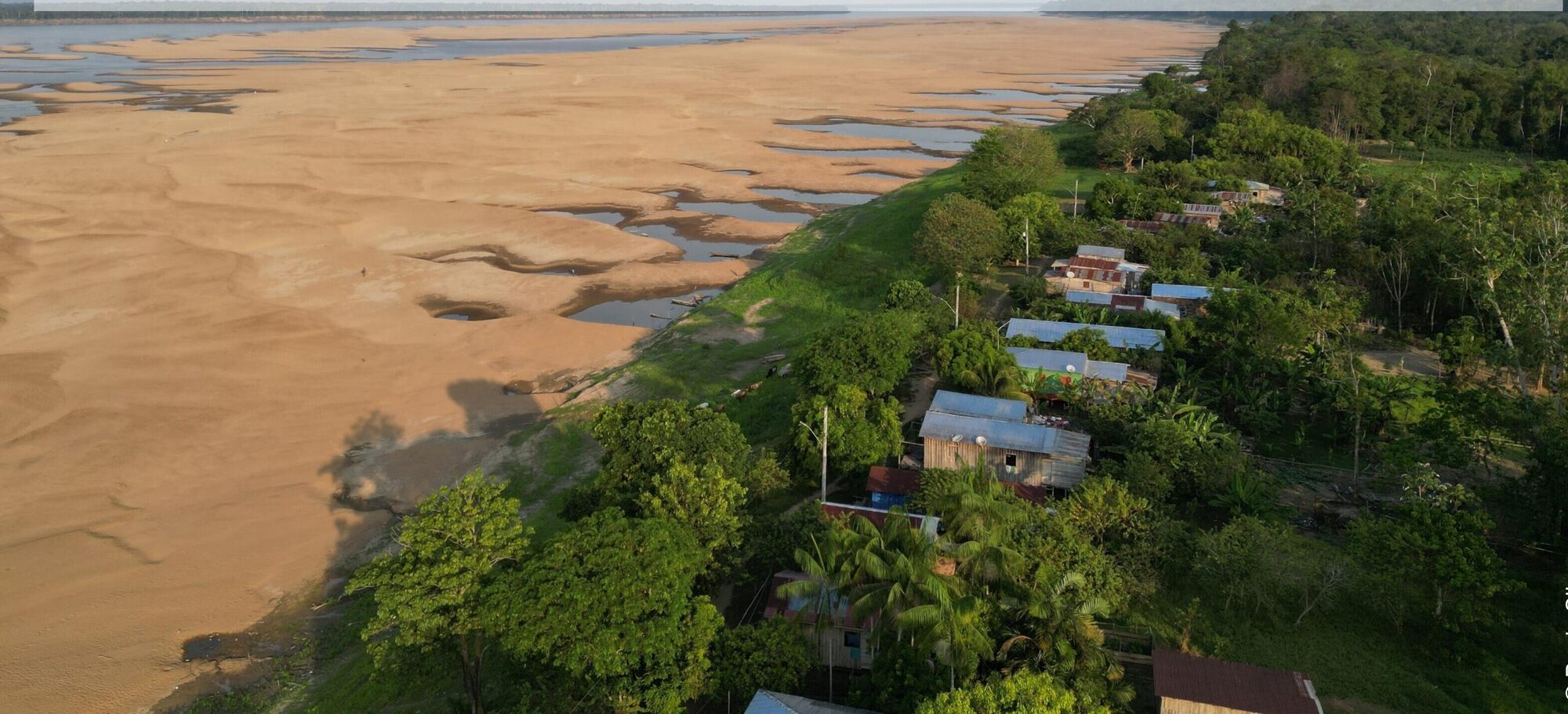 Vista aérea de lago seco com bancos de areia e uma comunidade na margem, cercada de floresta